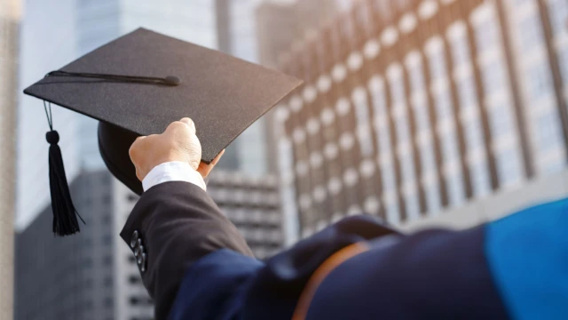 A stock photo showing the outstretched arm of a graduate. He is wearing a funny rope and holds his academic cap in his end. In the background are tall, generic office buildings.