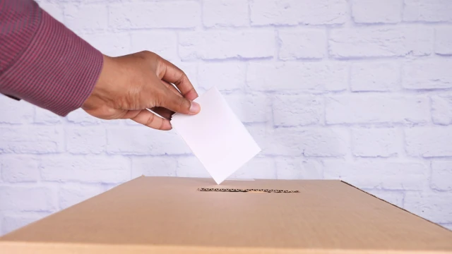 A stock photo of a hand putting a piece of paper in a ballot box made out of cardboard.
