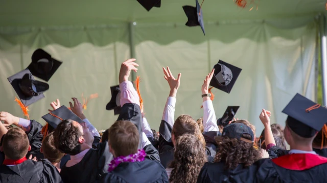 A stock photo of a crowd with posh university students. The students are seen from the back and they are facing what looks like a stage. They are all wearing ropes, and most them are busy throwing funny square hats into the air.