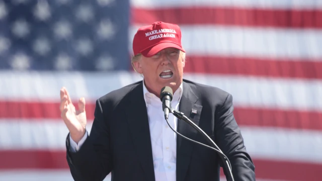 A photo of Donald Trump delivering a speech. He is wearing a red baseball cap with the text Make American Great Again and stands in front of a large American flag.