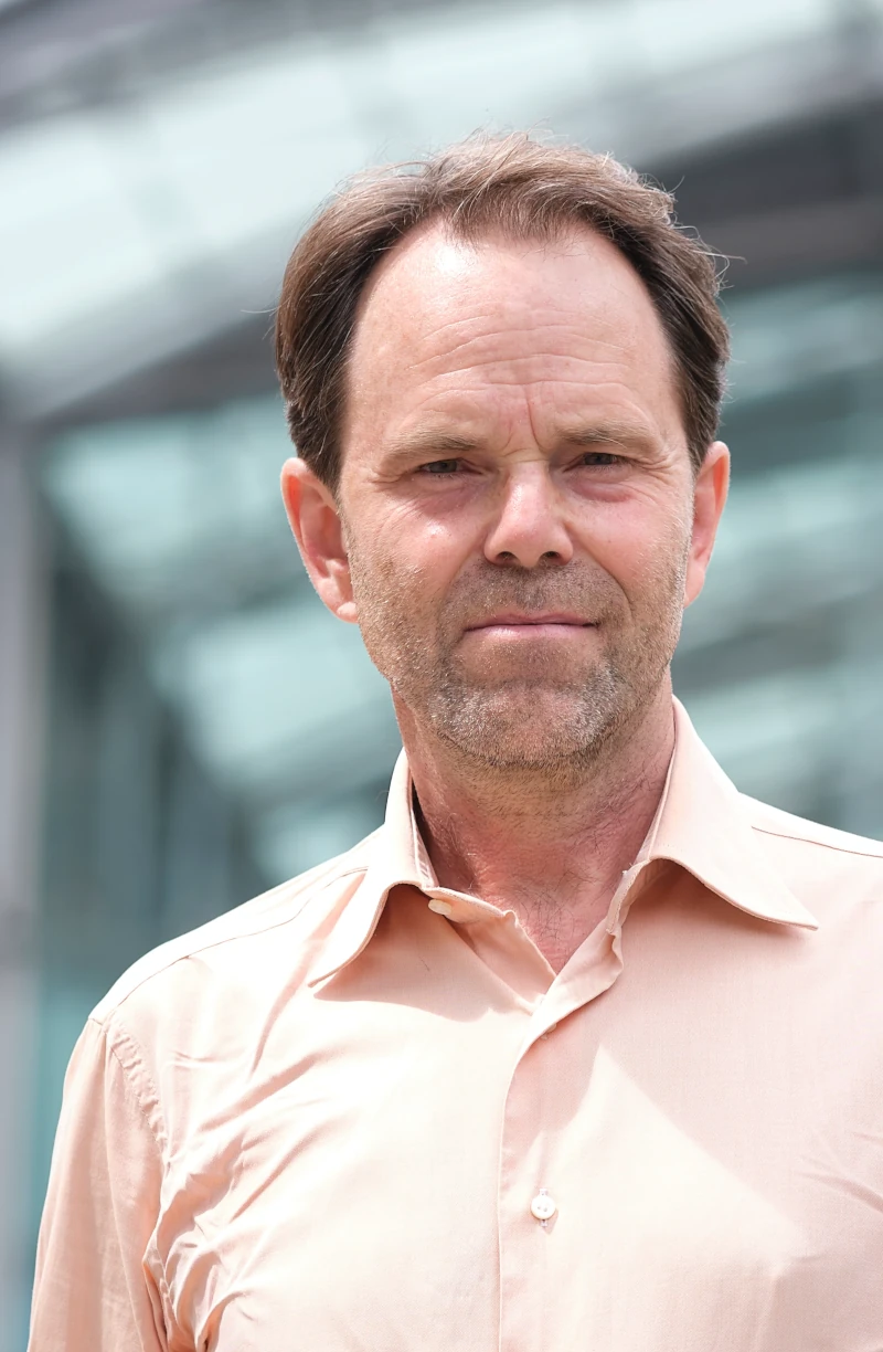 A photo of Rupert posing outside The Forum in Norwich. He is wearing a light shirt and is looking directly at the camera. The photo was captured from a slight low-angle perspective, which means you only see the large glass roof of The Forum in the background.